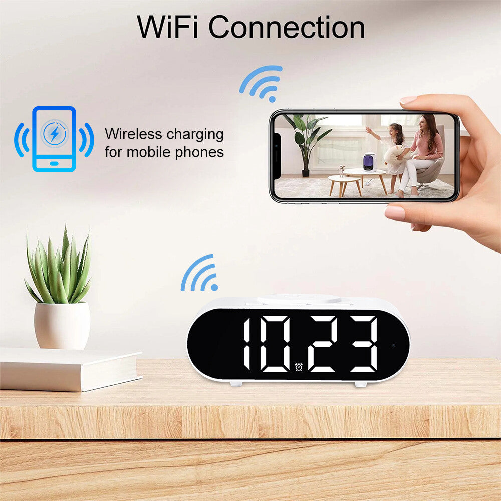 The Digital Clock Charger SpyCam with a large display sits on a wooden surface next to a plant, offering wireless charging and WiFi surveillance as the phone shows the live camera feed.