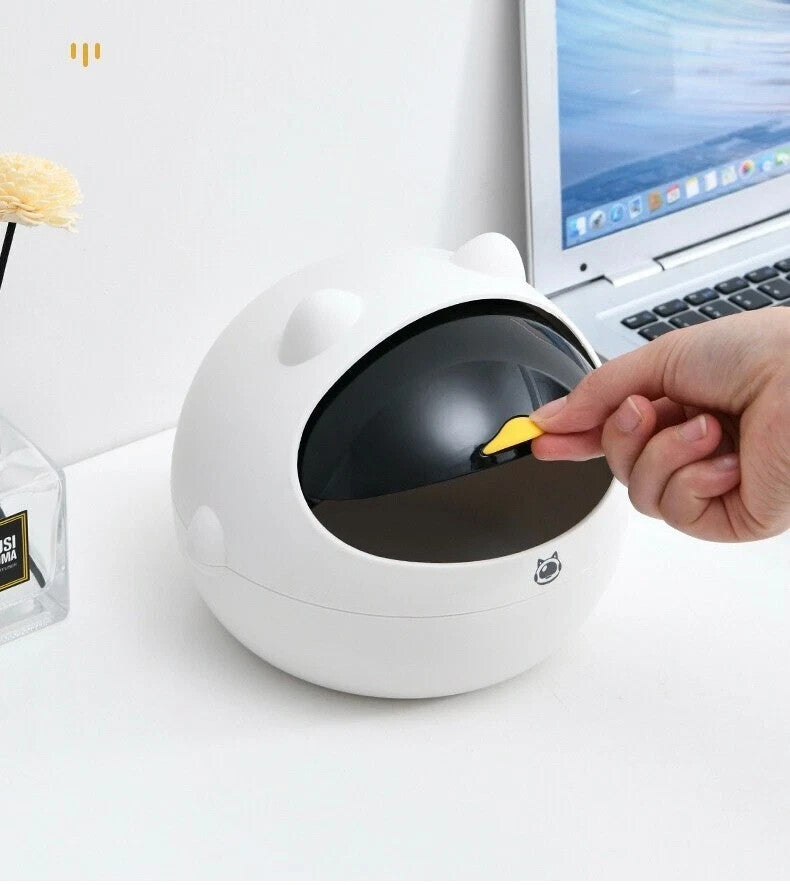 A hand puts the Cute Display SpyCam, a small yellow gadget, into a bear-shaped white container with a black opening beside a laptop—an adorable and discreet home surveillance accessory.