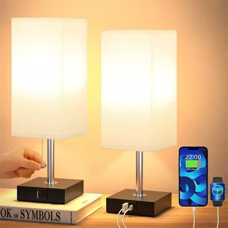 Two Desk Lamp Charging SpyCams with white rectangular shades on black bases, one lit. A smartphone and smartwatch charge via USB ports on the James Bond-inspired bases.
