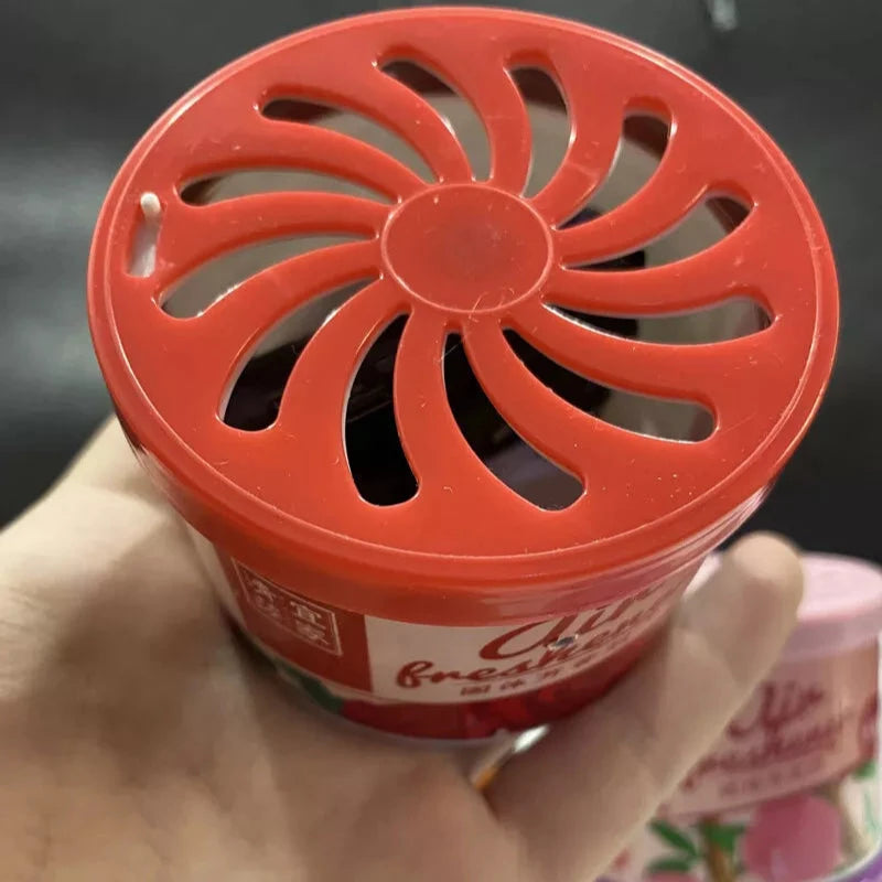A hand holding a Fake Decorative Display SpyCam, disguised as a red container with a vented lid labeled as an air freshener, subtly conceals the hidden camera. Another similar container is partially visible in the background. Perfect for high-tech surveillance needs.
