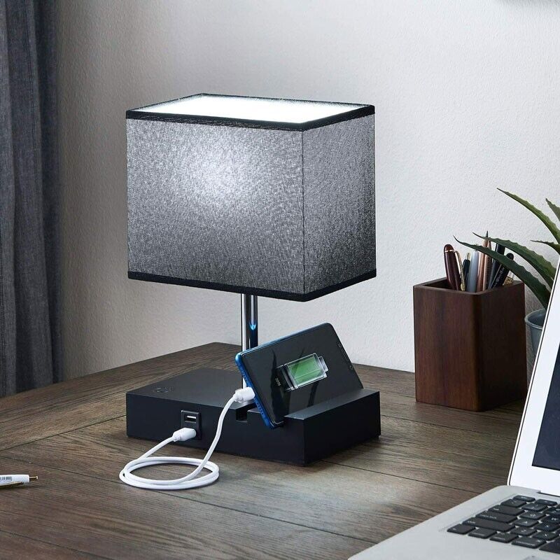 The Desk Lamp Charger SpyCam, featuring a rectangular shade on a wooden desk, discreetly integrates surveillance with a phone charging dock, USB cable, and smartphone. A plant and pen holder are visible in the background.