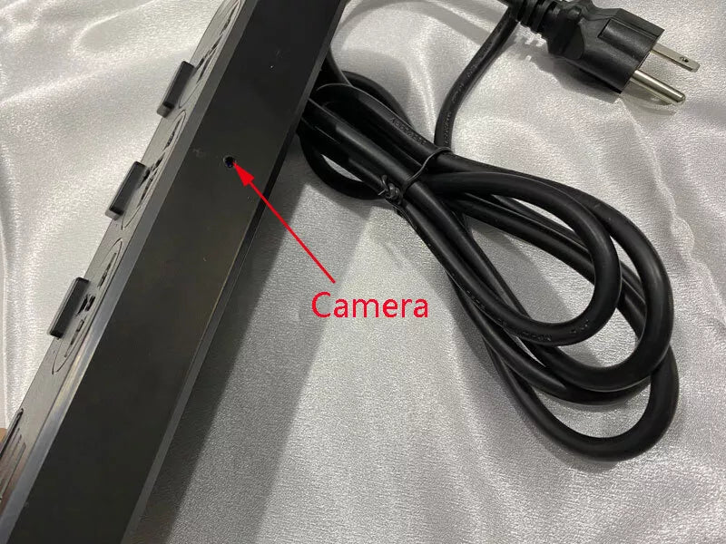 A close-up of the Power Strip SpyCam featuring multiple outlets, a black cord, and a three-prong plug. A red arrow highlights a small hole marked "Camera" on the side, indicating its covert surveillance capabilities with 1080P HD video quality.
