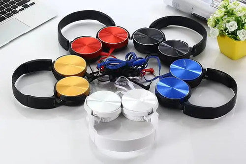 Six pairs of Wired Bass Headphones in red, yellow, blue, black, white, and silver are arranged in a circle on a white surface near a laptop and a yellow flowerpot, highlighting their ergonomic design.