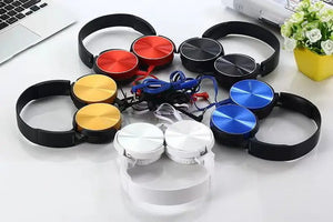 Six pairs of Wired Bass Headphones in red, yellow, blue, black, white, and silver are arranged in a circle on a white surface near a laptop and a yellow flowerpot, highlighting their ergonomic design.