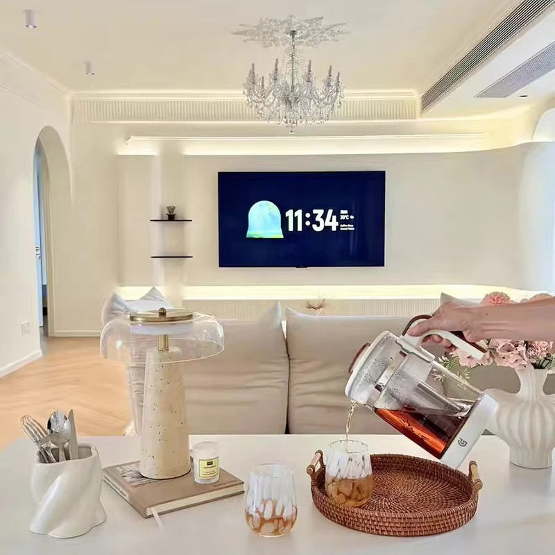 Modern living room with chandelier, wall-mounted TV displaying 11:34, white sofa, and coffee being poured into glasses on a table adorned with flowers and a Mushroom Desk Lamp in the foreground.