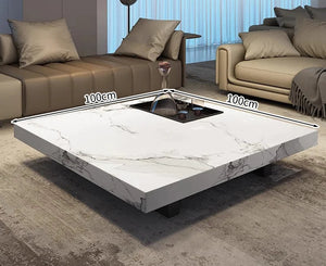 The Modern Luxe Coffee Table features a square marble top, black legs, and a recessed center compartment. Measuring 100 cm per side, it’s ideal for modern interiors and is displayed with sofas and cushions in a living room setting.