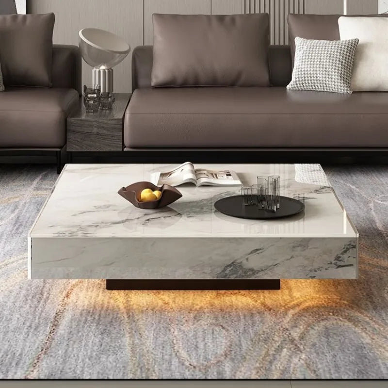 The Modern Luxe Coffee Table, featuring a square marble design with a tray of glasses, fruit bowl, and magazines, sits between two brown sofas in a modern living room styled for contemporary interiors.