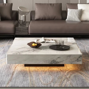 The Modern Luxe Coffee Table, featuring a square marble design with a tray of glasses, fruit bowl, and magazines, sits between two brown sofas in a modern living room styled for contemporary interiors.