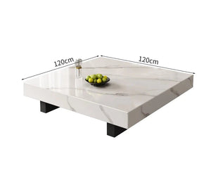 The Modern Luxe Coffee Table is a square marble design with black legs (120cm x 120cm), ideal for modern interiors. Styled with a small dish of green apples and a bottle.