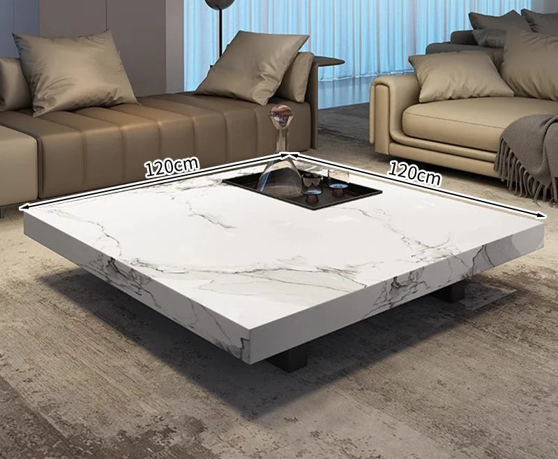 The Modern Luxe Coffee Table, a square white marble piece with a black center compartment (120cm x 120cm), sits on a rug between two sofas—an elegant focal point for modern interiors.