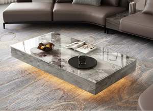 The Modern Luxe Coffee Table, rectangular and underlit, sits in front of a brown sofa on a patterned rug, styled with a glass of water, magazines, and a decorative fruit bowl—ideal for modern interiors.