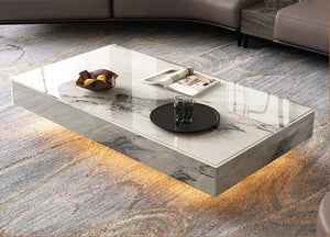 The Modern Luxe Coffee Table, featuring a rectangular glass top, rests on a patterned rug and displays a fruit bowl, tray with two glasses, and magazines. Warm under-lighting makes it ideal for stylish modern interiors.