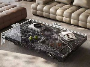 The Modern Luxe Coffee Table, a rectangular black marble piece (140x70x34cm) adorned with books, a tray, a decanter, glasses, and green apples, stands before a beige sofa—ideal for modern interiors.