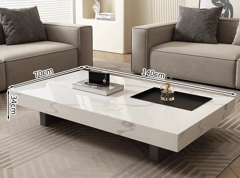 The Modern Luxe Coffee Table, a rectangular white minimalist design with black legs (140cm x 70cm x 34cm), sits between two gray sofas in a modern living room—ideal for contemporary interiors.