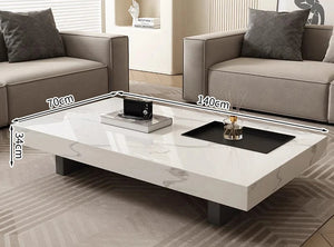 The Modern Luxe Coffee Table, a rectangular white minimalist design with black legs (140cm x 70cm x 34cm), sits between two gray sofas in a modern living room—ideal for contemporary interiors.