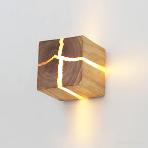 The Wood Cube Wall Light, designed in a contemporary cube shape and finished with a natural wood grain, showcases glowing cut-out lines and is elegantly mounted on a white wall.