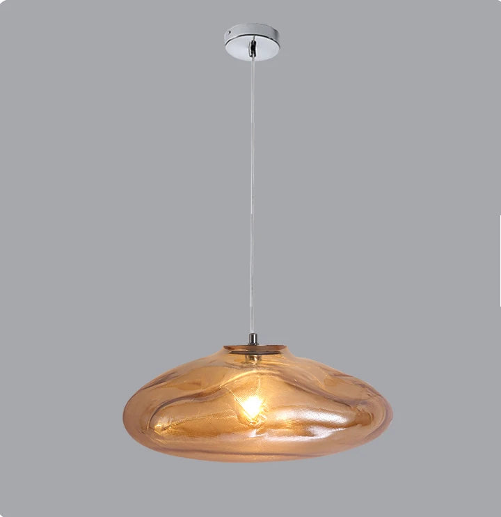 The Luxe Cloud Glass Pendant Light features a modern, sculptural design with a round amber-tinted glass shade suspended from a white ceiling mount against a plain gray backdrop.