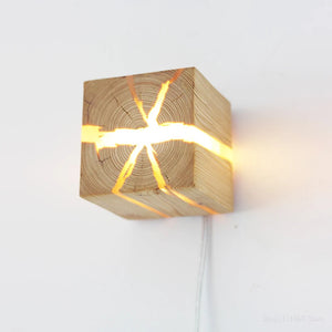 The Wood Cube Wall Light boasts a contemporary design, with its natural wood grain revealing glowing cracks that enhance warmth and elegance when installed on a white wall.