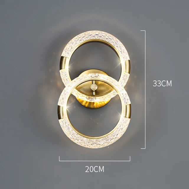 The Elegant Ring Wall Sconce is a luxurious wall light fixture featuring two interlocking illuminated rings, with dimensions of 33 cm in height and 20 cm in width. The gold-colored rings and base add an elegant touch to any space.