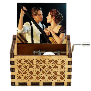 The Wooden Music Box is a hand-cranked music box with a carved pattern, showcasing an image of a man and woman in vintage clothing inside, with one of them turning the crank handle on the side.