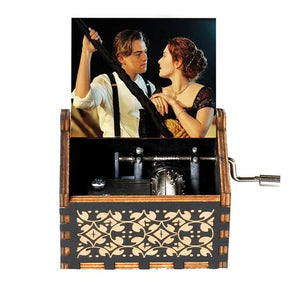 The Wooden Music Box is a handmade, hand-cranked treasure adorned with an image of a man and woman in a romantic scene. It features intricate designs on the sides and offers a glimpse of its inner mechanism, making it perfect for playing classic tunes.