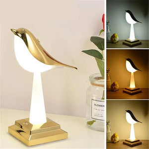 A Bird Table Night Light featuring a golden body and white base is displayed on a tabletop. Nearby, a flower and minimalist decor item enhance the setting. Smaller images showcase the night light in various tones, casting a warm glow.