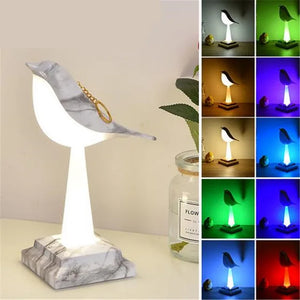 A Bird Table Night Light set on a marble base is illuminated, with an array of photos displayed next to it, each showcasing its contemporary design in various colors. A jar labeled "Flower" and a plant complement the scene, highlighting the lamp's elegant craftsmanship.