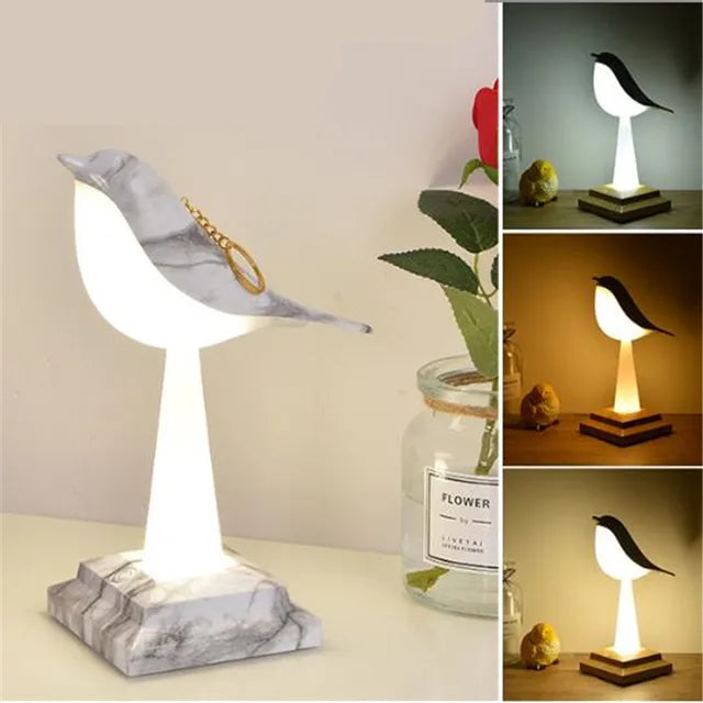 The Bird Table Night Light, showcasing elegant craftsmanship and contemporary design, features a bird-shaped body with a marble base. It includes three additional images displaying the light illuminated in various lighting conditions, positioned beside a jar and a rose.