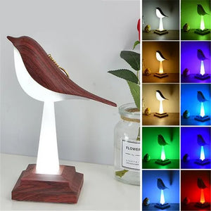 The Bird Table Night Light, showcasing elegant craftsmanship, is a wooden bird-shaped lamp on a stand, casting its glow next to a potted plant. On the right side, the same lamp with contemporary design emits various colors including yellow, blue, purple, red, green, and white.