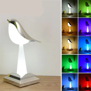 The Bird Table Night Light, a contemporary lamp, sits gracefully on a wooden table, showcasing elegant craftsmanship. It emits light in multiple colors arranged in a grid with nine variations: white, red, yellow, blue, green, and purple.