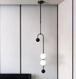 The Minimalist Line Light is a modern pendant featuring a sleek mounting system with a metal rod and three sculptural bulbs—two white, one black—hanging elegantly against a panel wall.