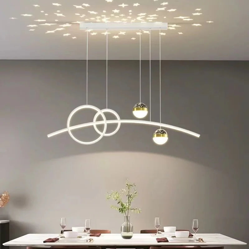 A modern dining room showcases a Stellar Pendant Light, blending circular and linear designs to cast celestial accents on the ceiling. A table for four is elegantly set with plants and wine glasses, creating an inviting ambiance.