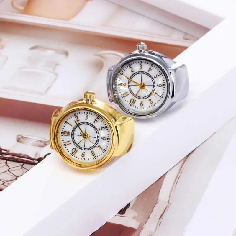 Two small Vintage Ring Watches, one gold and one silver, rest on a white geometric surface. Each showcases a pearlescent dial, blending personalized luxury with timeless vintage style.