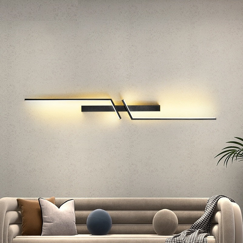 The Minimalist Wall Sconce, a modern geometric wall-mounted LED fixture, illuminates a beige wall above a beige sofa with mixed cushions, with a plant seen on the right.