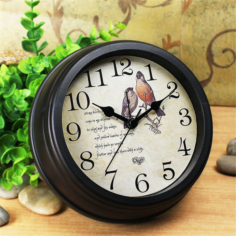 Analog wall clock with a vintage bird illustration on its face, displaying the time approximately at ten minutes past ten, placed on a wooden surface with greenery and pebbles around it. This piece